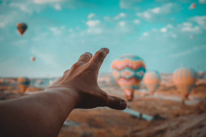 A forced perspective photo of a persons hand reaching toward hot air balloons using orange and teal color scheme 