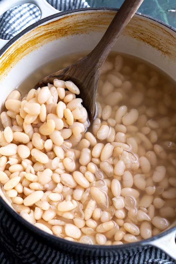 wooden spoon scooping cooked white beans in white dutch oven on striped linen